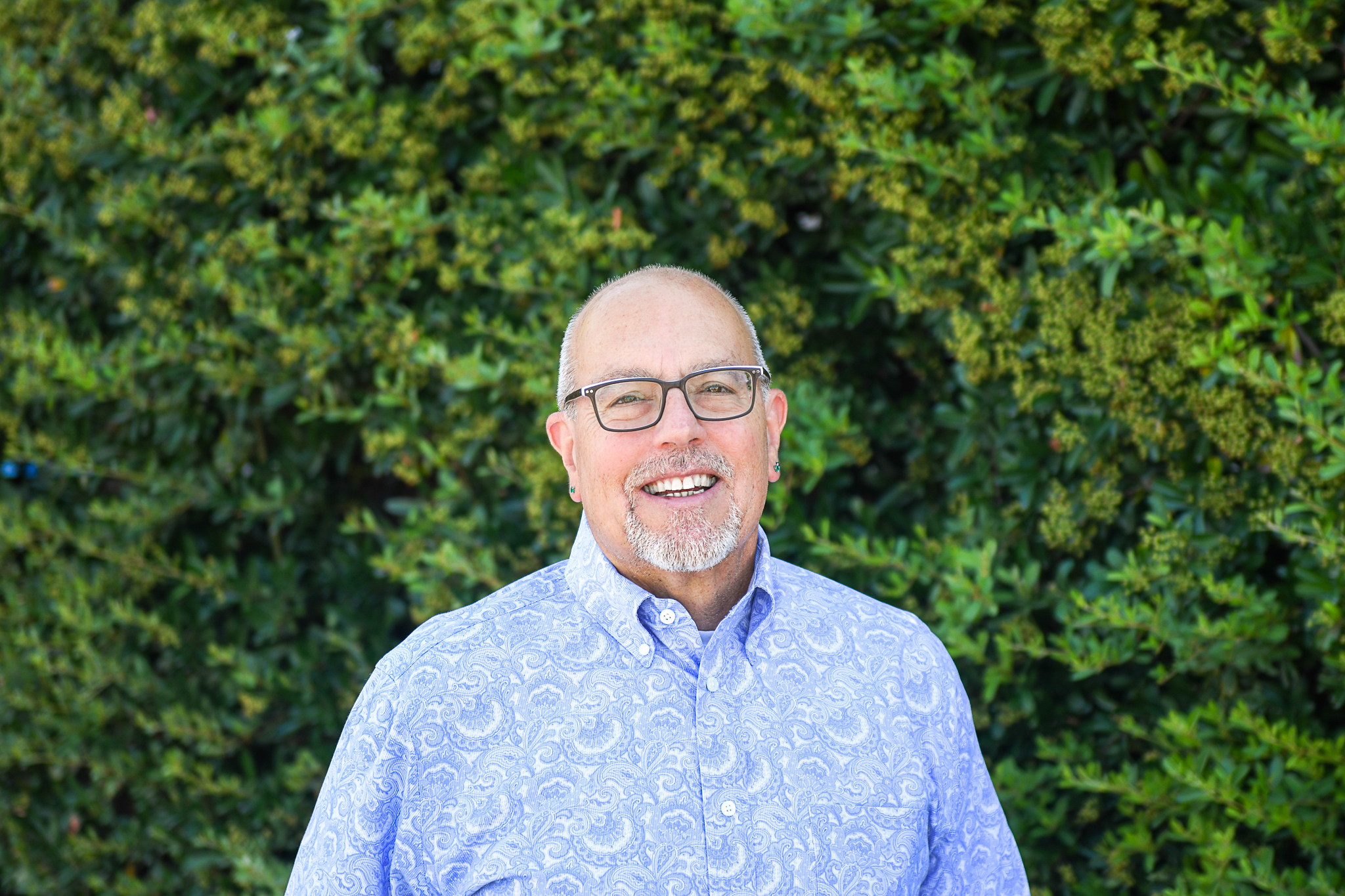Craig Galati smiling in front of a green hedge